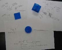 A photograph shows three blue plastic pieces of varying shapes and sizes (two square, one circular) with three half-sheets of white paper nearby containing sketches of each, with parts identified.