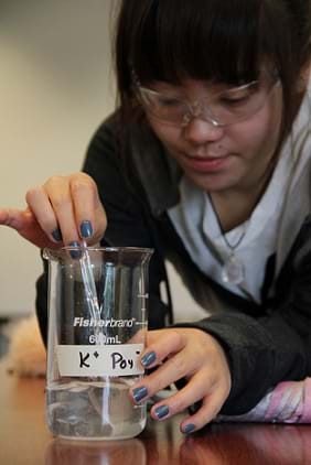 A young girl wearing safety glasses stirs a cloudy liquid in a glass beaker.