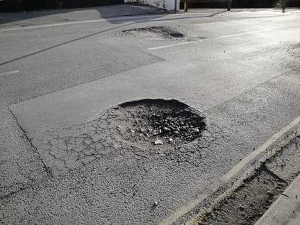 A photograph shows a generally smooth asphalt road with two potholes. One is circular and large. The other is smaller and more of an asphalt indentation. 