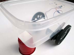A photograph shows a clear rectangular plastic food container serving as a vehicle frame with two drive servos taped to the back of it with two attached wheels, and a small cup holding up the front of the container.