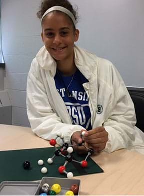 A photograph of a girl using an atom model set to construct a glucose molecule on a classroom table.