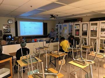 A teacher is shown presenting the PowerPoint slides while students are taking notes and listening. 