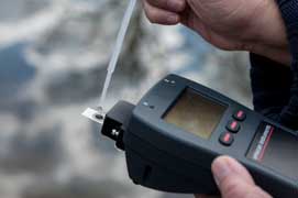 Photo shows a drop of liquid being placed on a sensor plate connected to a handheld plastic device with a small display screen and control buttons. 