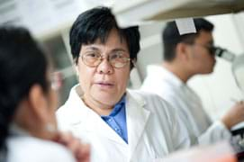 Photo shows a woman in a white lab coat talking to another person. Behind her, a man looks into a microscope.