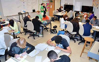 A photograph shows a classroom of teens, four at a table, with every one of them with heads down, intently working with papers, pencils and cookies in front of them (cookie mining).