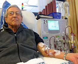 A photograph shows a man leaning back into a chair with his left arm resting on its armrest. Tubes inserted into his arm are connected to a nearby machine with a monitor and dials.