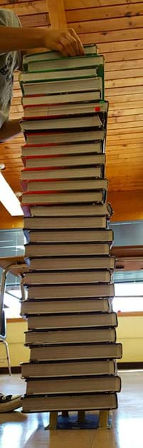 A photograph shows a student stacking the 27th same-size hardback textbook on top of a student-created structural protein model.