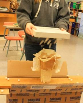 A photograph shows a student holding a flat rectangular-shaped box over a student-designed transport protein model with mini marshmallows coming out of the box.