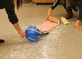 A photograph shows two students holding a balloon and piece of cardboard over scattered piles of shredded paper on the floor.
