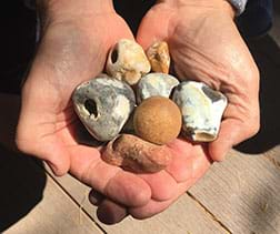 A photograph shows two cupped hands held together and holding about seven small, smooth and roughly round rocks of varying colors—white, gray, brown, orange.