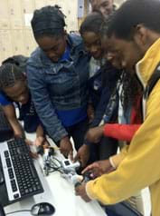 Five teenagers huddle around two robots being directed to pull apart from each other.