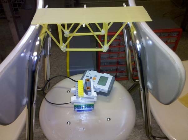 A photo shows a spaghetti truss bridge placed upside down and spanning across the chair backs of two chairs positioned back-to-back. Between the two chairs and directly beneath the center of the upside down bridge is a stool on which the LEGO brick connected to an ultrasonic sensor rests, with the sensor pointing directly upwards at the center of the bridge span.