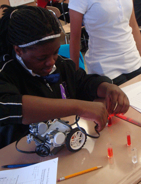 A photograph shows a student at a table using a LEGO robot to shine light from a small device into orange liquid in a vial.