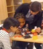 A photograph shows two students and a teacher focuses intently on an activity at a table.