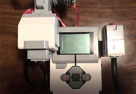 Photo shows a LEGO hand-held plastic device with a display window connected to a servomotor, a smaller box-shaped plastic device. Two gray triangular-shaped buttons are identified with arrows as "1." The servomotor is identified with an arrow as "2."