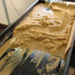 A photograph shows a long, black plastic tray with six-inch sides filled with beige sand. At one end, a small stream of water pours in, traveling down the table through a meandering canyon in the sand.