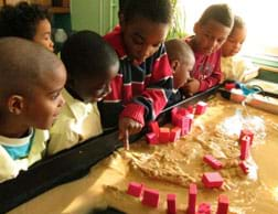 A photograph shows seven young students standing along a sand-filled erosion table, engaged in noticing the flow of the water and which model buildings remain standing and which have collapsed due to erosion.