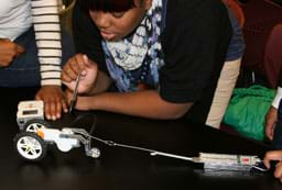 Students huddle at a table with a handheld computer device and a robot that has two large back wheels and two small front wheels and is attached by line to a small metal box.