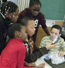 Five students crowd around a table as one boy handles a plastic bag of ice and water and another boy reads a hand-held display.