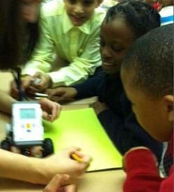 Students gather around a desktop robot, using pencils to mark on the paper under the robot.