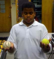 A boy stands with his eyes closed and arms outstretched, holding a baseball in one hand and a tennis ball in the other.