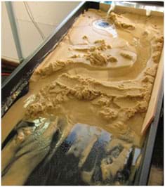 A photograph shows a long, black plastic tray with six-inch rims filled with beige sand. At the far end, a stream of water flows in, traveling down the table through a meandering canyon in the sand.
