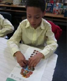 Photo shows a student at a table placing pennies inside the outline of his foot, which was traced onto graph paper.