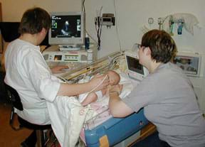 A photograph shows a person sitting at a computer and holding a wired device on the chest of an infant lying in a crib nearby; the baby's mother sits on the other side of the crib.