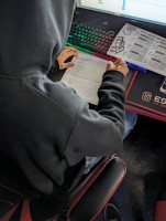 A photo showing a student wearing a dark hoodie seated at a computer station with a red-and-black esports-themed desk. The student is working on a printed worksheet titled "4 Input Logic Gate," using a pencil to complete a truth table, referencing a logic gates handout while looking at a screen that displays gate logic information.