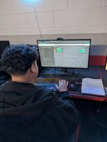 A photo of a student sitting at a computer desk using Wokwi, a logic circuit simulator, to test a custom logic gate design. The student is wearing a dark hoodie and is focused on the monitor, which displays logic gates, input switches, and a code window. A worksheet labeled “Logic Gates III” sits on the desk, partially completed with drawn gates and truth table outputs.