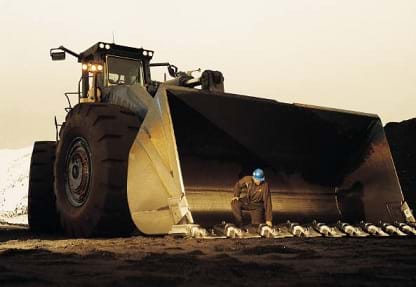Photo shows a man looking small inside the scoop bucket of a huge industrial front-end loader