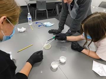 Students in personal protective equipment gather around a laboratory workspace and test bacteria samples in agar dishes.
