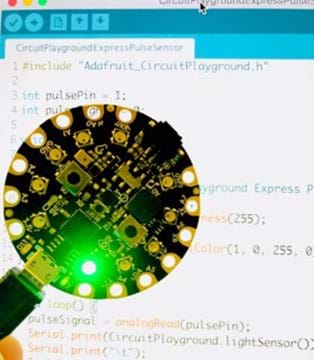 A photograph shows a Circuit Playground Express board and behind it is some programming code on a computer.