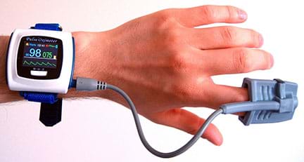 Picture of a wrist mounted pulse oximeter. The sensor is attached to the finger, which in turn is attached to the device via a cable.
