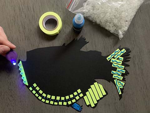 An aquatic organism cut-out on black cardstock expressing bioluminescence with fluorescent materials such as glow-in-the-dark tape, UV beads, and glow paint. 