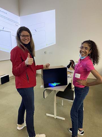 A student and teacher pose with their test animal. A black diorama cardboard box mimicking the deep ocean contains an aquatic organism cut-out modeling bioluminescence using fluorescent materials. A mini UV light is shining on the prototype.