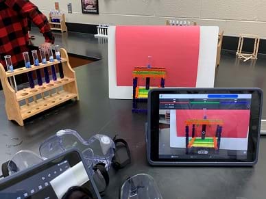 Experimental setup on the table. A test tube holder holds five test tubes containing blue liquid; another test tube is held in the sample holder which sits in front of a red piece of paper; an iPad open to a camera-based app is pointed at the sample holder; safety glasses sit on the table.