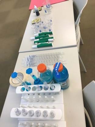 Materials laid out on a table, including test tube holders and test tubes, two bottles of blue Gatorade, masking tape, a pile of pipettes, beakers, a small bottle of blue dye, and red construction paper.