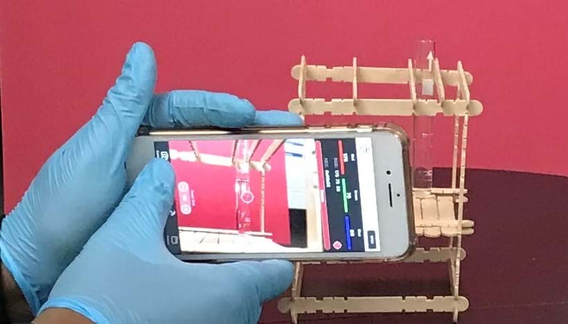 A student holds up a cell phone with a camera-based app open to measure the absorbance of food dye sample which is sitting in a sample holder.