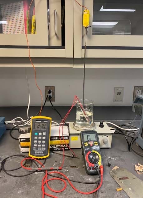 A photo showing the full set up for the resistance tests. A thermometer measures the temperature, a voltmeter measures the resistance, and a camera captures both screens to get the most accurate reading for the data collection.