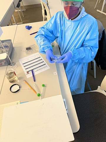 A student in personal protective equipment (PPE) checks the seal on a petri dish in a class laboratory.