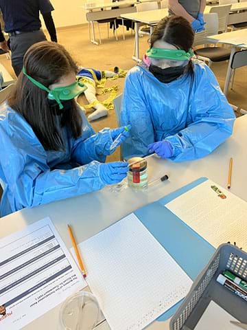Two students in personal protective equipment (PPE) test a lab sample for contamination in a classroom setting. 