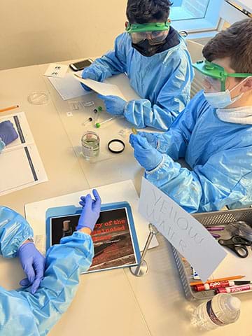 A group of three students in personal protective equipment (PPE) analyze a sample and take notes at a lab station.