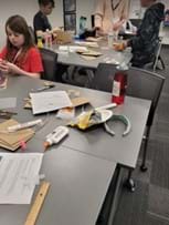 A photo of students gathering in groups at desks to work on prototypes using piping and classroom materials. 
