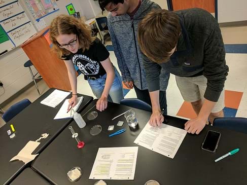 Three students in a lab are measuring salt to prepare different salinity concentrations using graduated cylinders, salt, and bottles of distilled water as they record their information and follow the lab’s instructions.