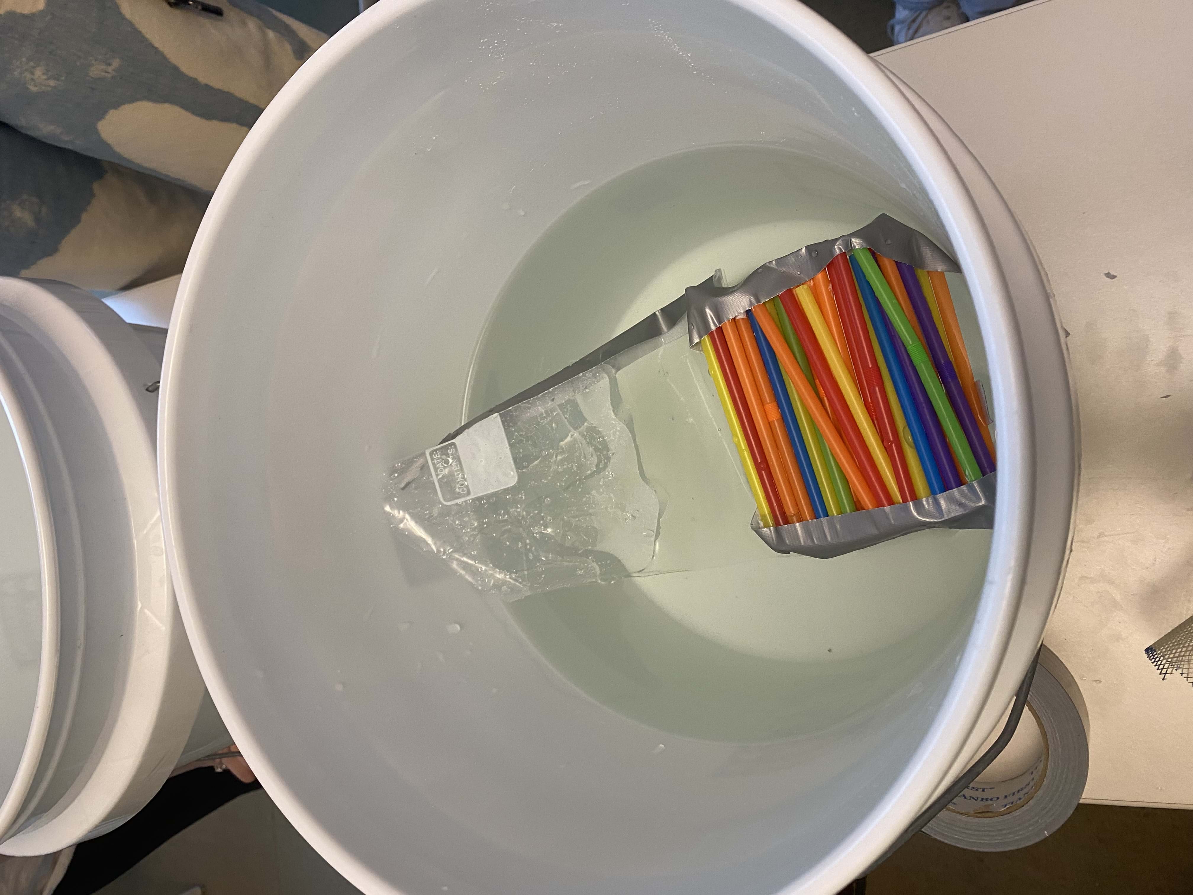 A white bucket that is filled halfway with tap water contains a prototype. The prototype is mostly made of straws that are duct taped together at the ends and attached to a plastic bag full of air. 