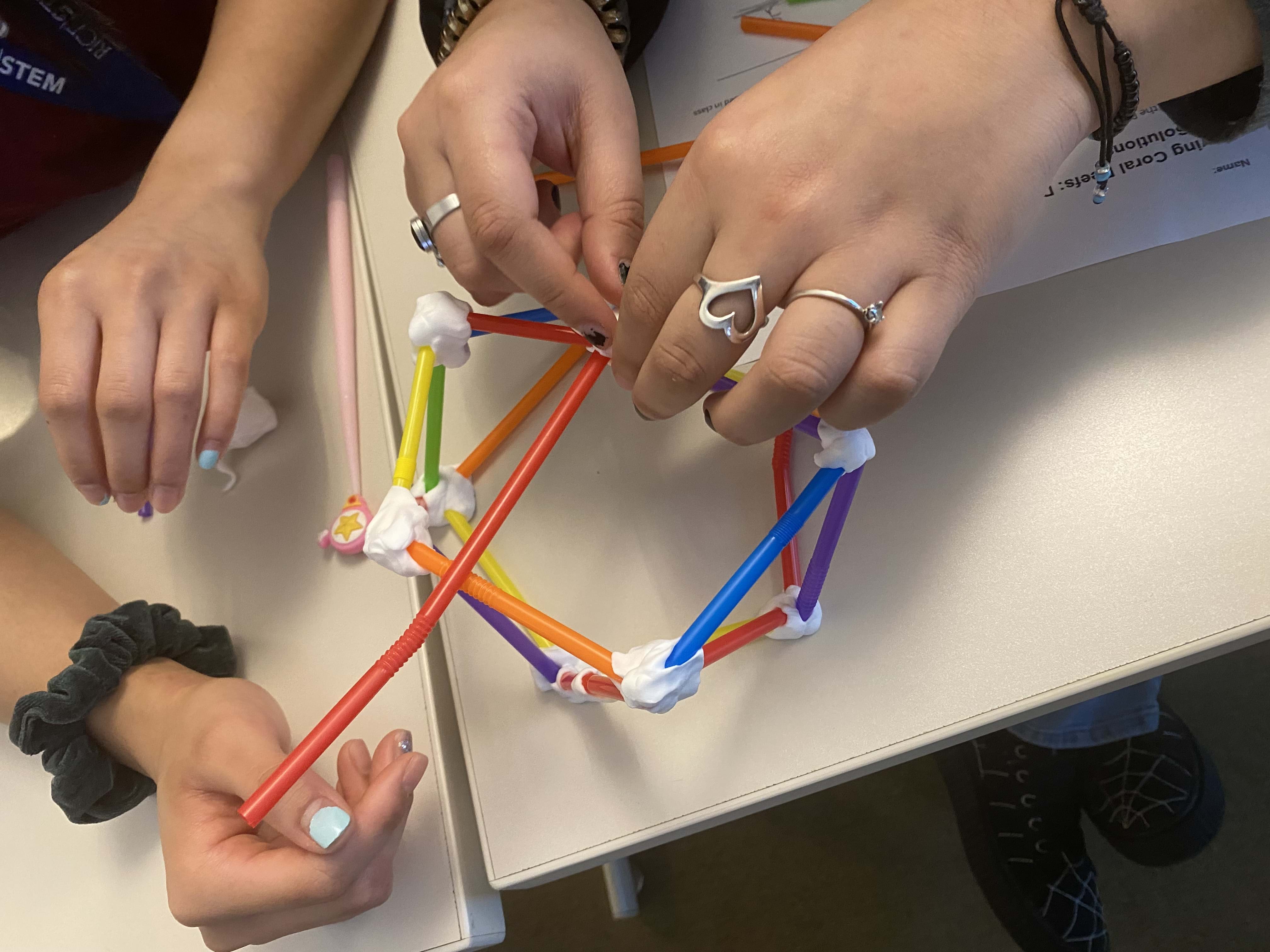 Two sets of hands are shown above a desk putting together pieces of straw using putty to create a ball shape as part of the original design for their first prototype.
