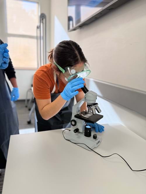 A photo showing a student in protective gear viewing a microplastics solution sample under a microscope. 
