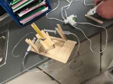 A photo showing a flat base constructed of wide popsicle sticks with a short popsicle stick tower in the center. On the tower are 4 wide popsicles put together as a platform that can rotate as the syringe pushes against it. The syringes are attached to the platform using popsicle sticks. When you push one, it causes the other syringe to extend out, pushing and moving the platform.