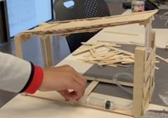 A photo showing a bridge constructed from popsicle sticks, with a rectangular platform as the base and a rectangular platform as the top/bridge portion. The top platform is held up in each corner using 2 wide popsicle sticks glued vertically to form a corner. The front of the bridge has a wall between 2 corners that is holding one syringe and the plastic tubing. The second syringe is attached to the base using zip ties. When the syringe on the base is pressed, it causes the syringe attached to the front wall to push up vertically and raise the top platform. The platform is lowered by pulling the syringe attached to the bottom base. 